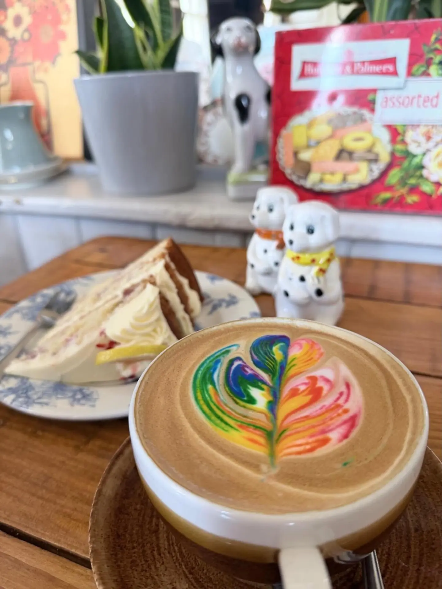 Coffee with rainbow latte art beside a slice of cake